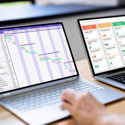 A project manager uses two laptop devices to manage Gantt charts and lean lab metrics, illustrating digital project management