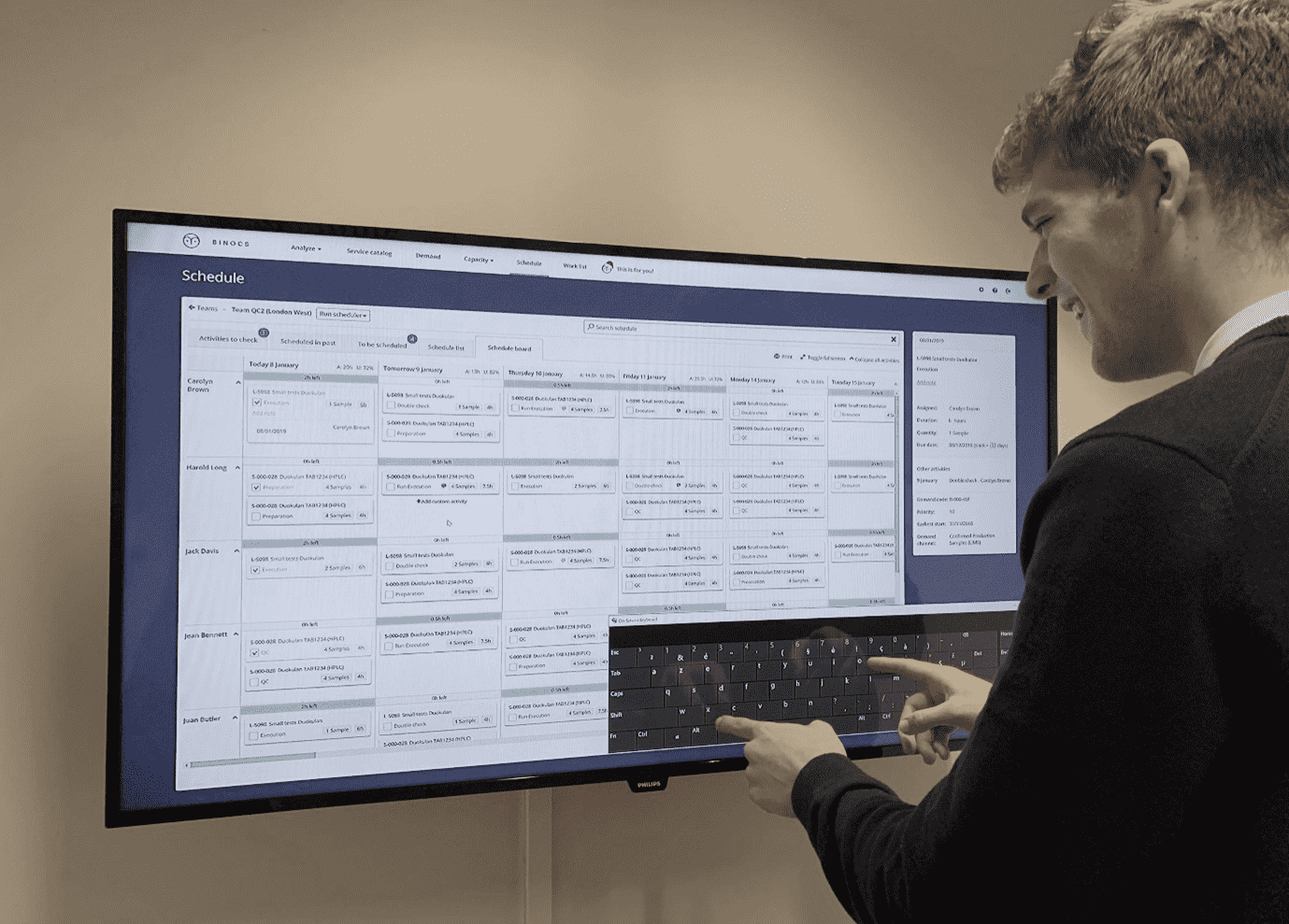 A man interacts with the Binocs schedule board via a large touchscreen display installed in a lab. This is a great example of how digital is changing lean labs