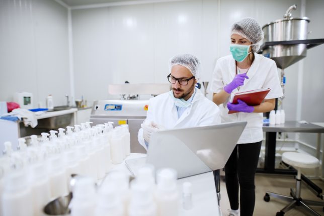 Scientist doing research in cosmetic factory. Next to him assistant with folder in hands. On the desk bottles and laptop.