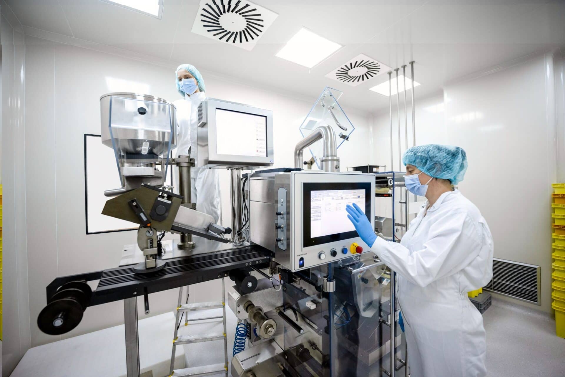 Female scientist in pharmaceutical industry seen standing in front of the screen on the very complex machine and taping on it while wearing protective white suit, blue gloves, a face mask and a cap during her day shift.